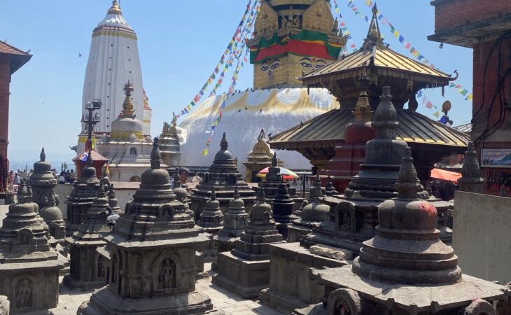 Multiple stone stupas in front of the Swayambhunath Stupa with colorful prayer flags.